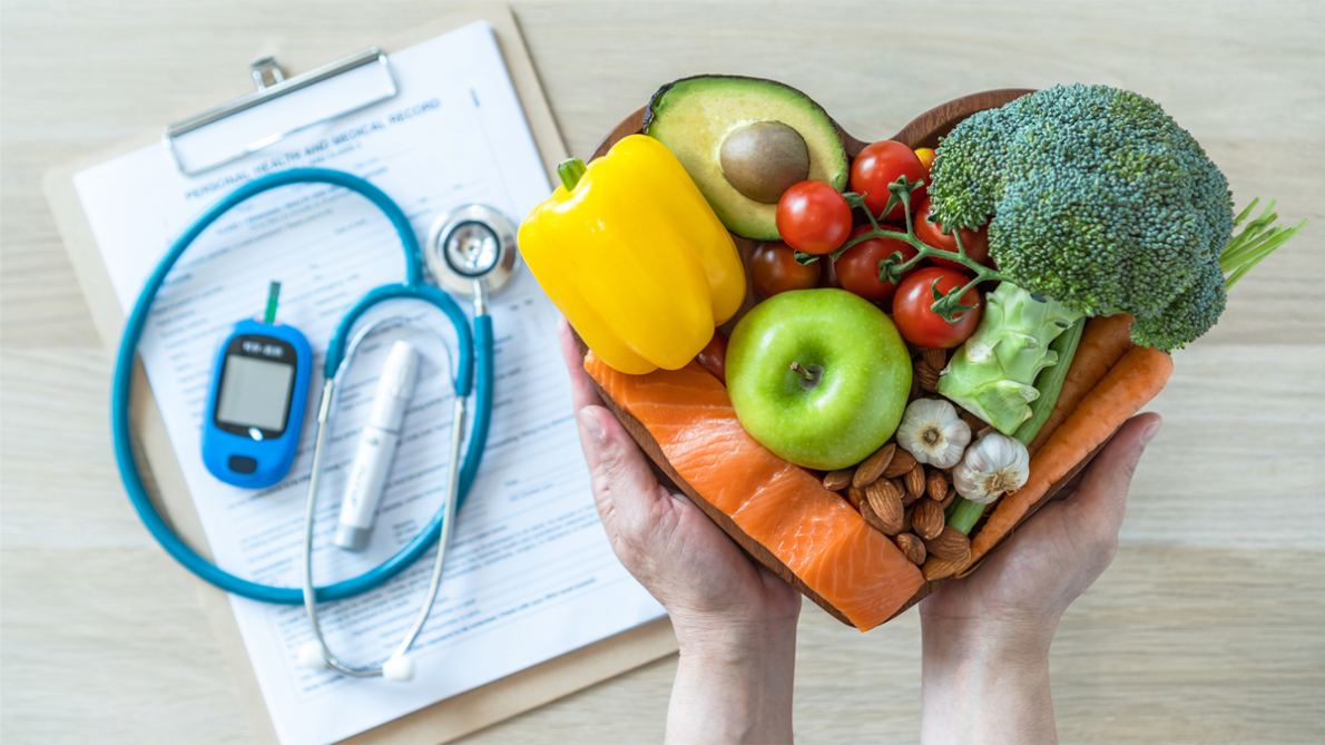 Hands holding a heart shaped wooden bowl filled with fruits, veggies and nuts. 