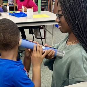 A student participating in the HPREP Junior program listens to a mentor's heart with a homemade stethoscope. 